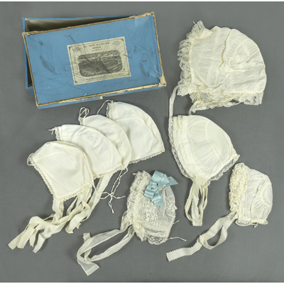 SLM 39030 1-9 - Åtta babymössor av bomull förvarade i fransk skokartong, från Ökna i Floda socken