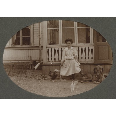 SLM P09-1506 - Kvinna framför hus med hund, katt och höna