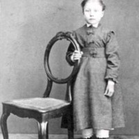 SLM M033637 - En okänd liten flicka står vid en stol.