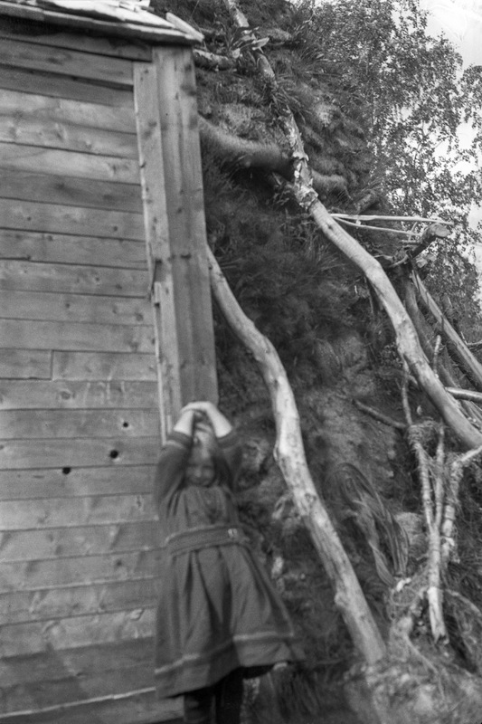 Flicka stående framför träbyggnad intill kåta.