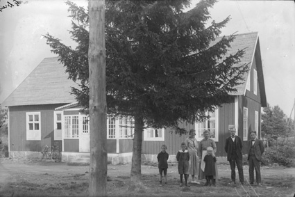 Familj (sju personer) framför trähus. Stor gran.