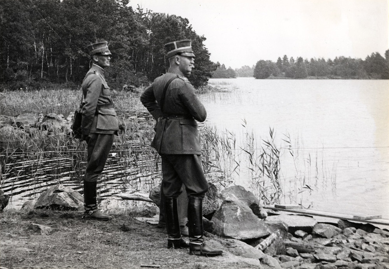 Tre militärbefäl står vid en sjö och samtalar.