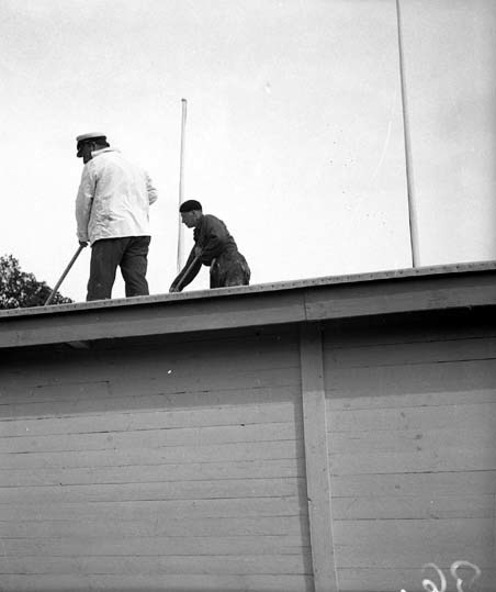 Två män som som är uppe på ett tak och målar, d...